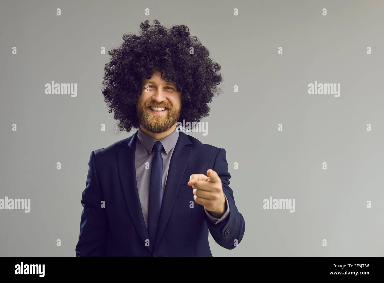 Toothy smiling businessman in curly toupee pointing to camera studio portrait Stock Photo