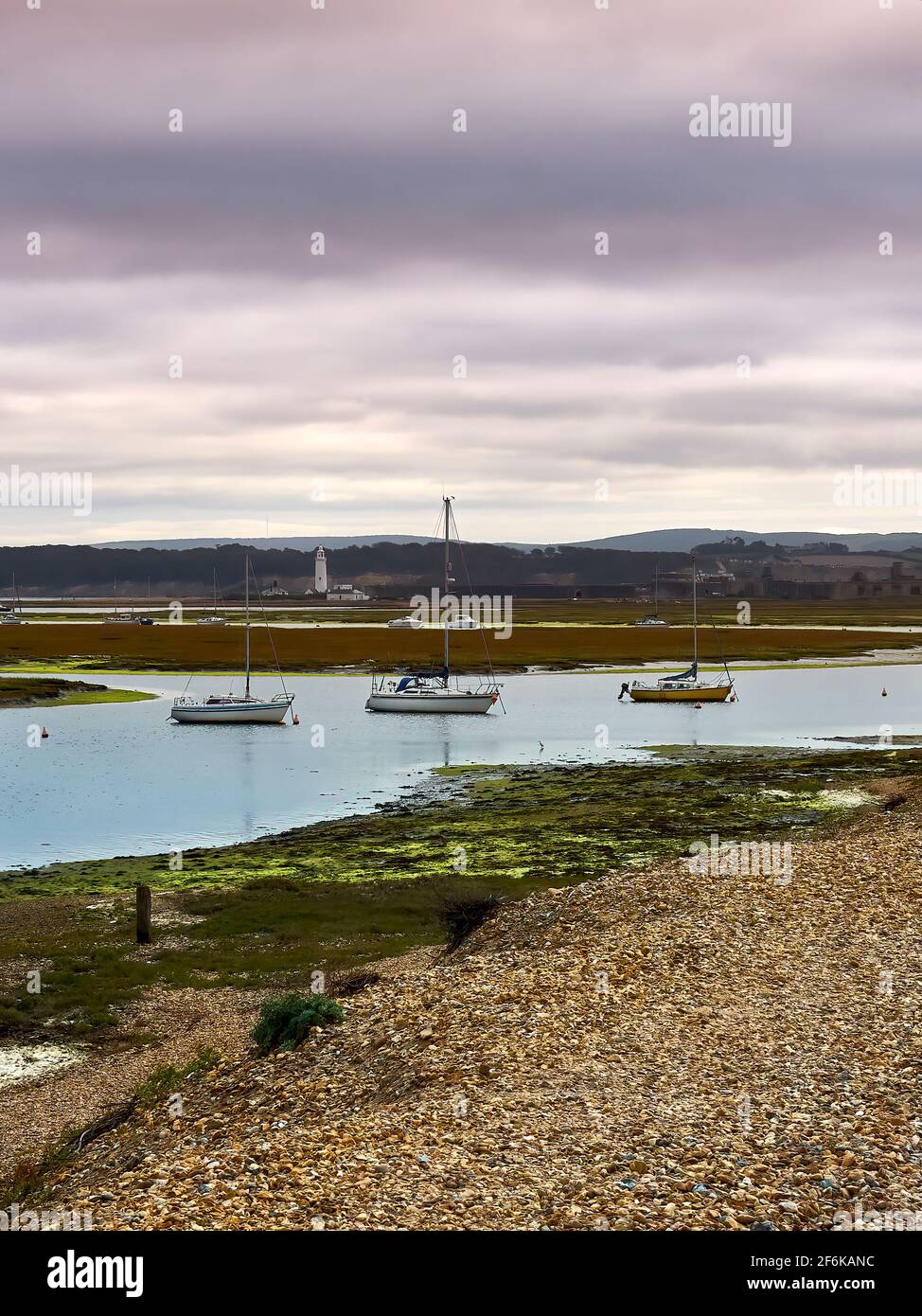 A shingle path next to a  creek running through marshland. Yachts are anchored on the creek with rolling hills, a lighthouse and a dramatic sky behind Stock Photo