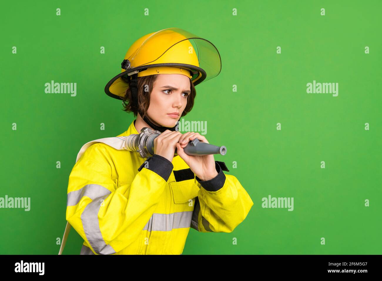 Photo of adorable serious firewoman dressed yellow uniform helmet holding fire hose looking empty space isolated green color background Stock Photo