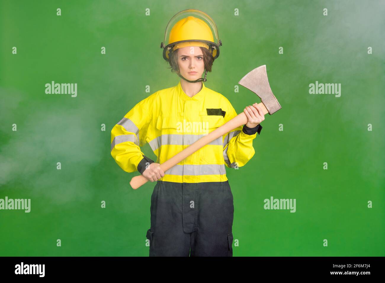 Photo of pretty serious young firewoman dressed yellow uniform helmet holding hatchet isolated green color background Stock Photo