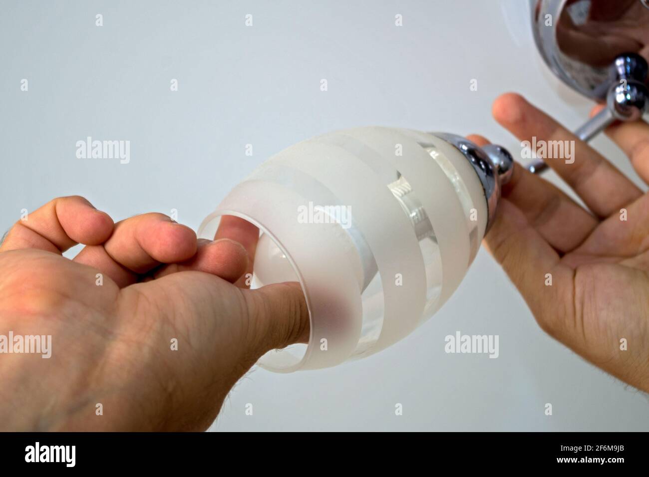 The left man's hand holds the arm of the chandelier the right hand screws or unscrews energy-saving ball-type lamp on white ceiling background. Concep Stock Photo