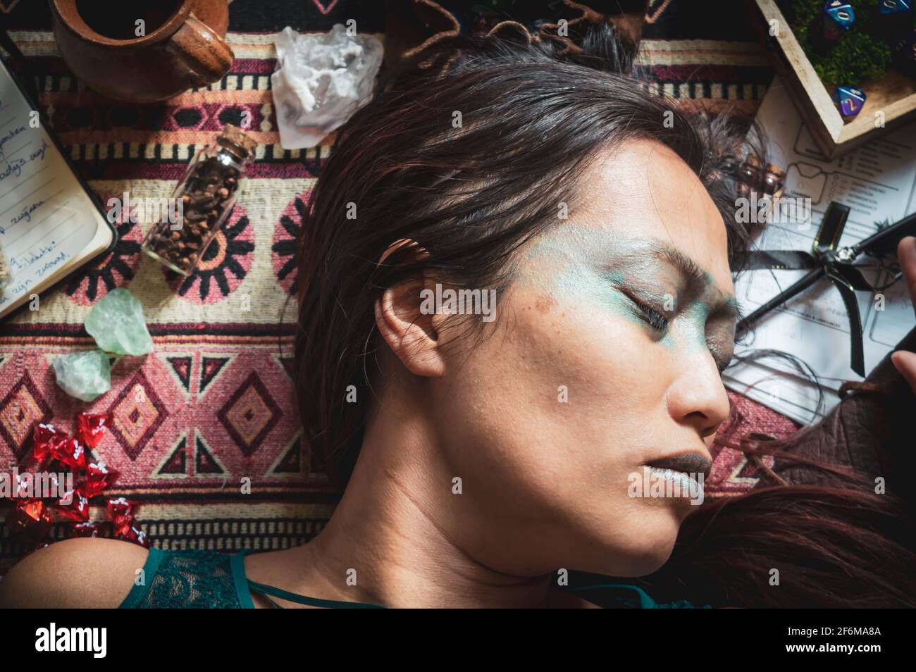 Portrait of a sleeping Indonesian woman surrounded by role-playing game attributes like dice, crystals pen and paper and, notebooks. Vintage edit. Stock Photo