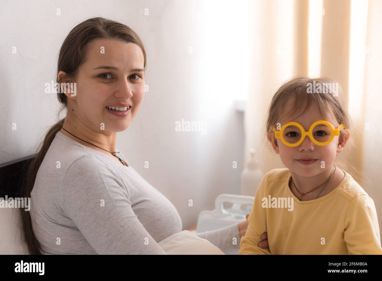 Cute little girl wearing uniform playing doctor or nurse with young mum or nanny in bedroom, checking mother throat, holding stethoscope, family Stock Photo