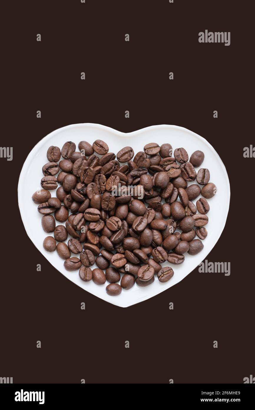 Coffee beans are poured on white plate in shape of heart. Brown background isolated. Stock Photo