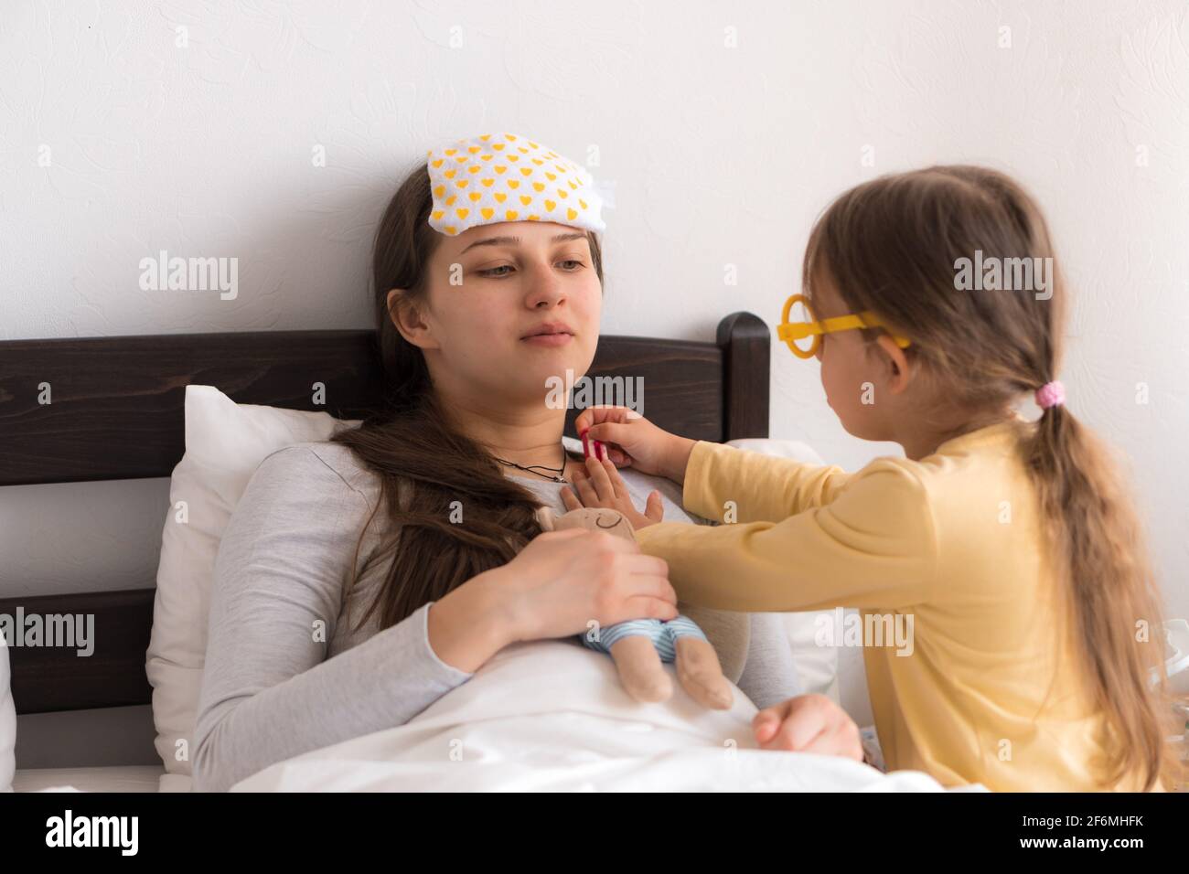 Cute little girl wearing uniform playing doctor or nurse with young mum or nanny in bedroom, checking mother throat, measures temperature, family Stock Photo