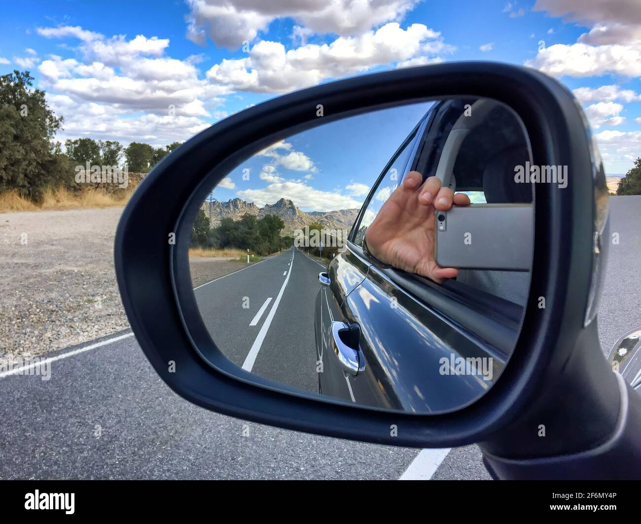 reflection in the rearview mirror of a car of a hand with a cell phone taking a picture of the reflection showing a road with a mountain landscape in Stock Photo