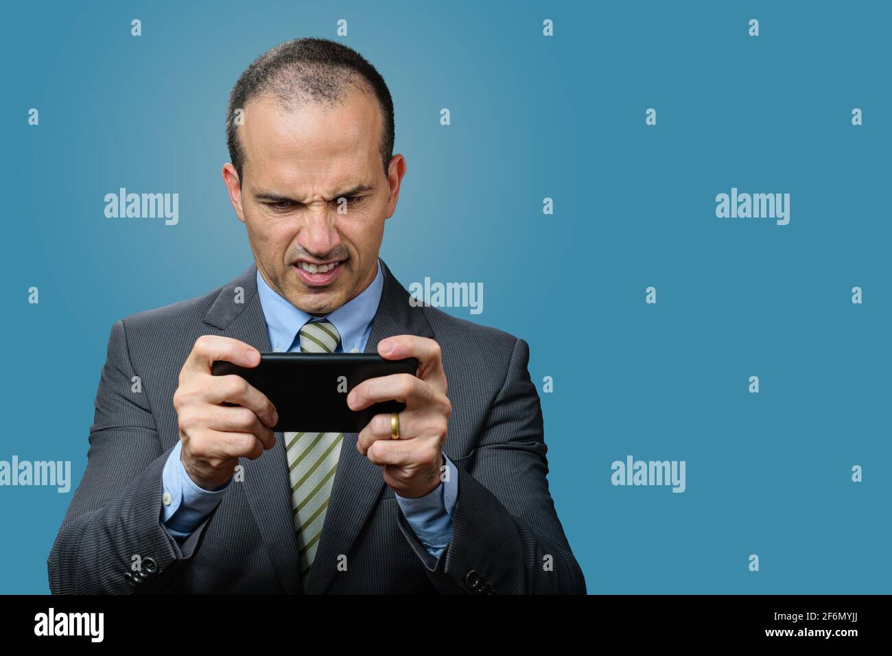 Mature man with suit and tie, playing on his smartphone and gritting his teeth. Blue background. Stock Photo