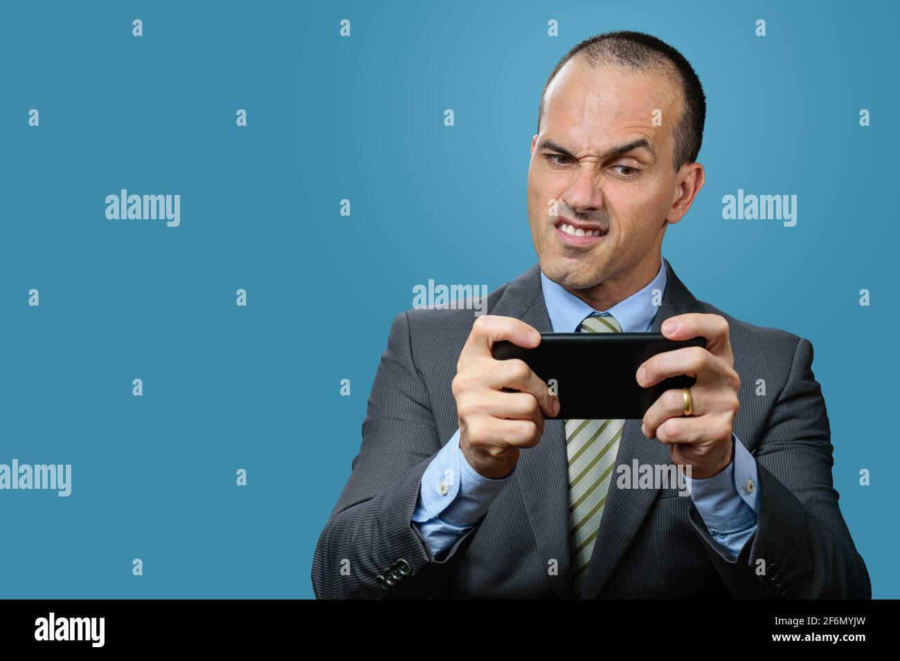 Mature man with suit and tie, playing on his smartphone and making a funny angry face. Blue background. Stock Photo