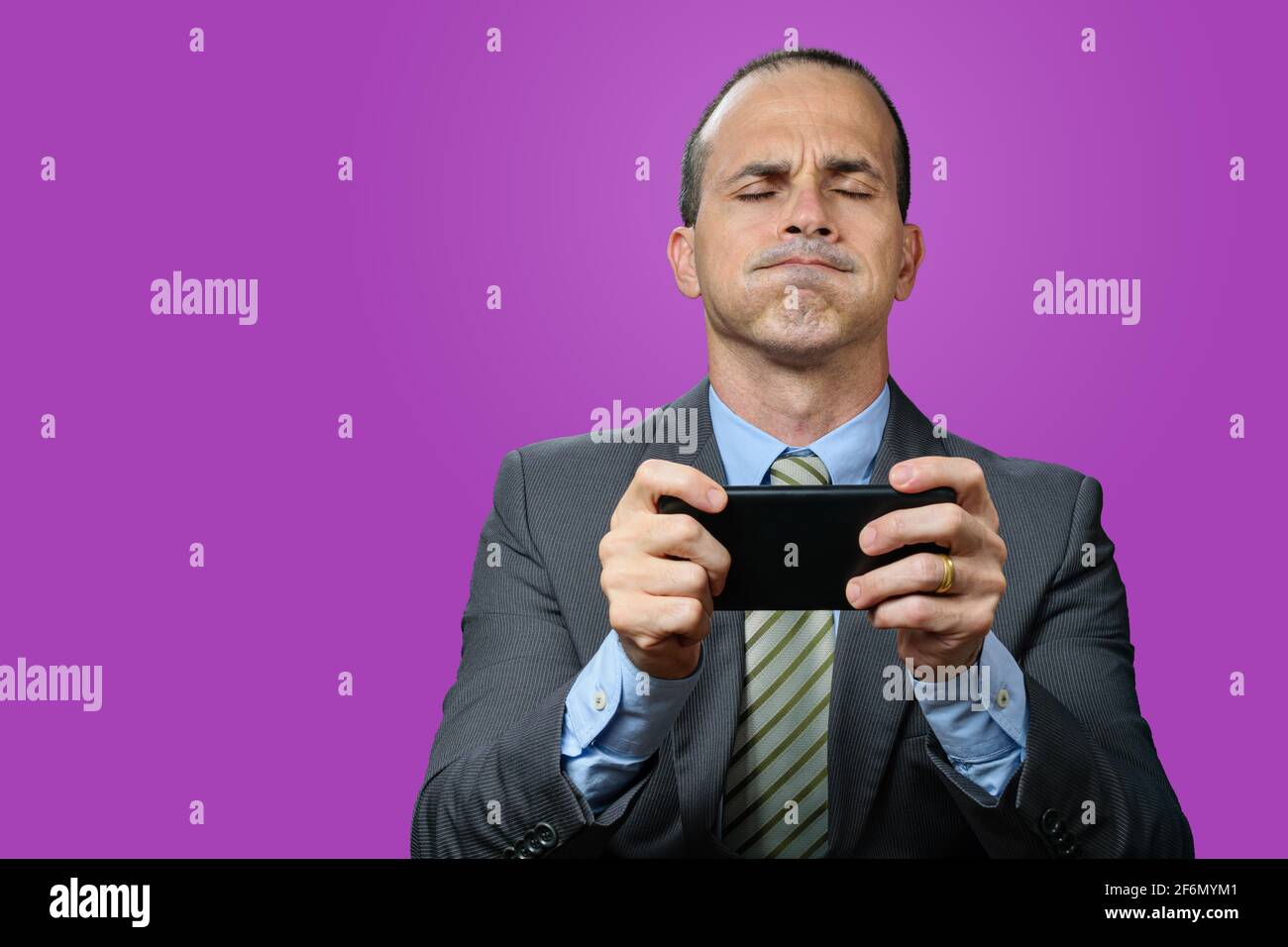 Mature man with suit and tie, holding his smartphone horizontally, breathing out through his nose and with his eyes closed. Purple background. Purple Stock Photo