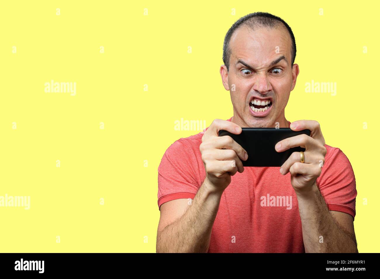 Mature man in casual clothing, with angry expression and holding smartphone horizontally. Yellow background. Stock Photo