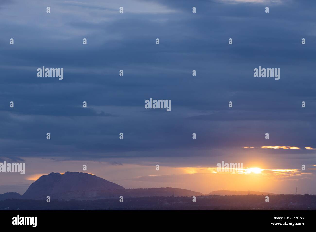 A Panoramic Landscape Silhouette of hills at the bottom with dark clouds in the horizon with sun peeping through them Stock Photo