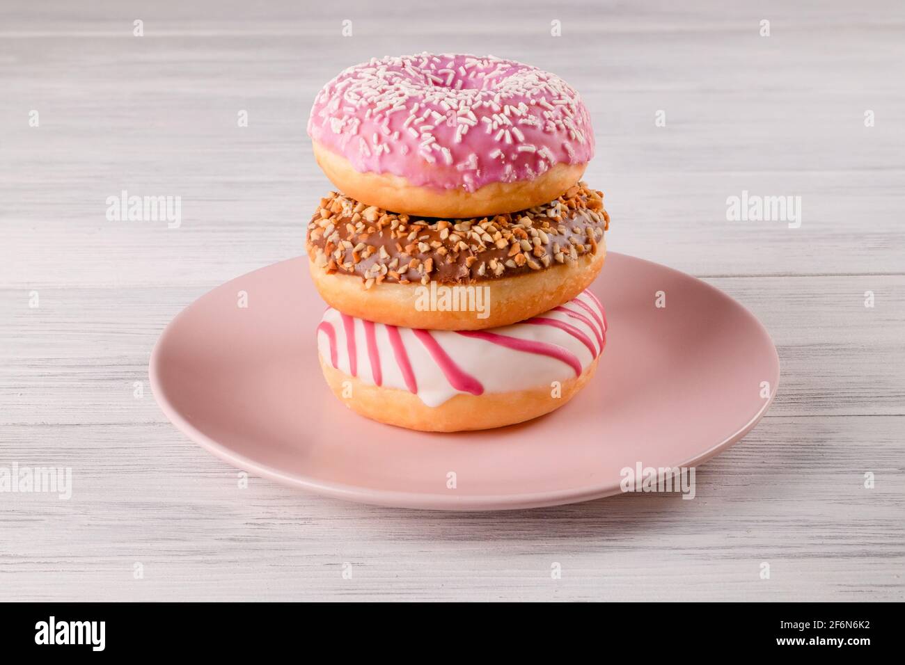 Three sugar-coated donuts are stacked on a pink ceramic plate on a light-colored wooden surface. Selective focus. Stock Photo