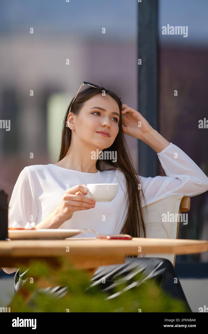 Dreamy young woman sitting on the terrace of the cafe and drinking ...