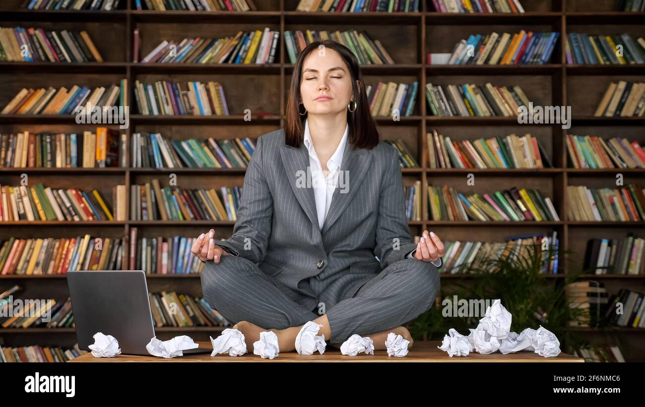Short haired businesswoman meditates sitting in yoga pose on wooden table among rumpled paper sheets and laptop against bookshelves close-up Stock Photo