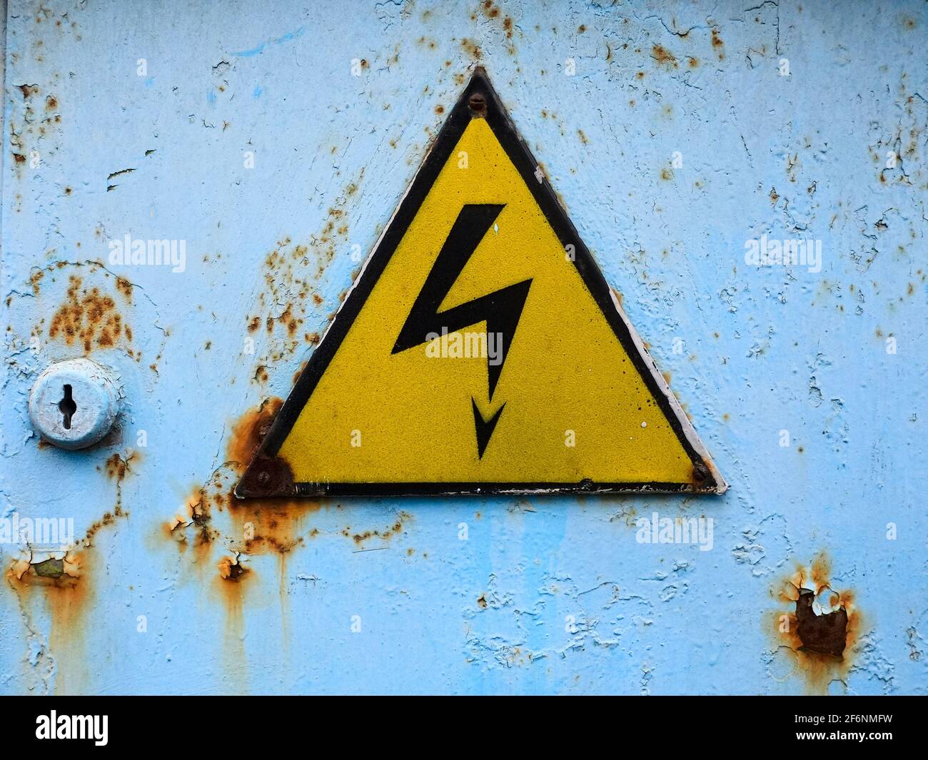 Old high voltage sign. Yellow triangle on a blue rusty door. Hazard danger symbol. Stock Photo