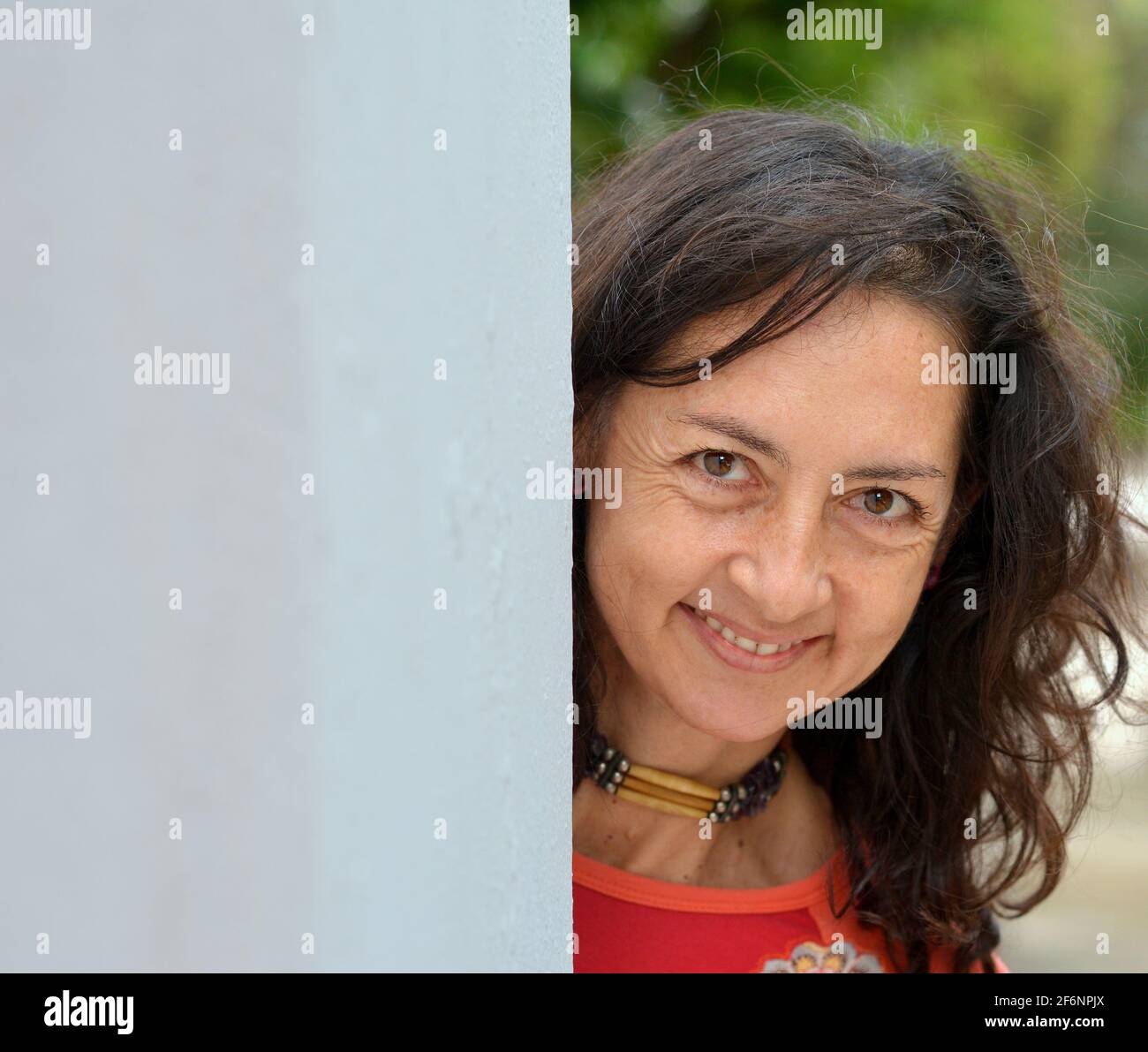 Charming mature Caucasian brunette woman with long hair peeks around a grey wall and makes a happy facial expression in front of a park background. Stock Photo