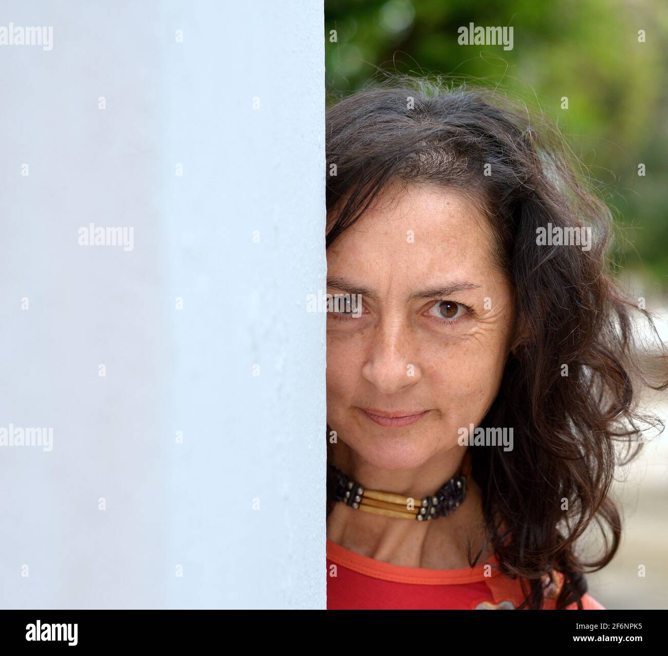 Serious mature pretty Caucasian woman with long hair peeks around a grey wall and makes a worried facial expression in front of a park background. Stock Photo