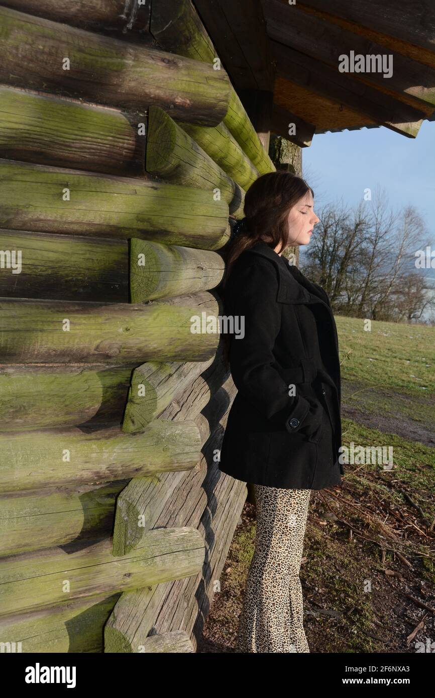 Young woman from the side, with black jacket, leaning against a wooden hut, with a serious expression on her face and closed eyes, enjoying the sun, i Stock Photo
