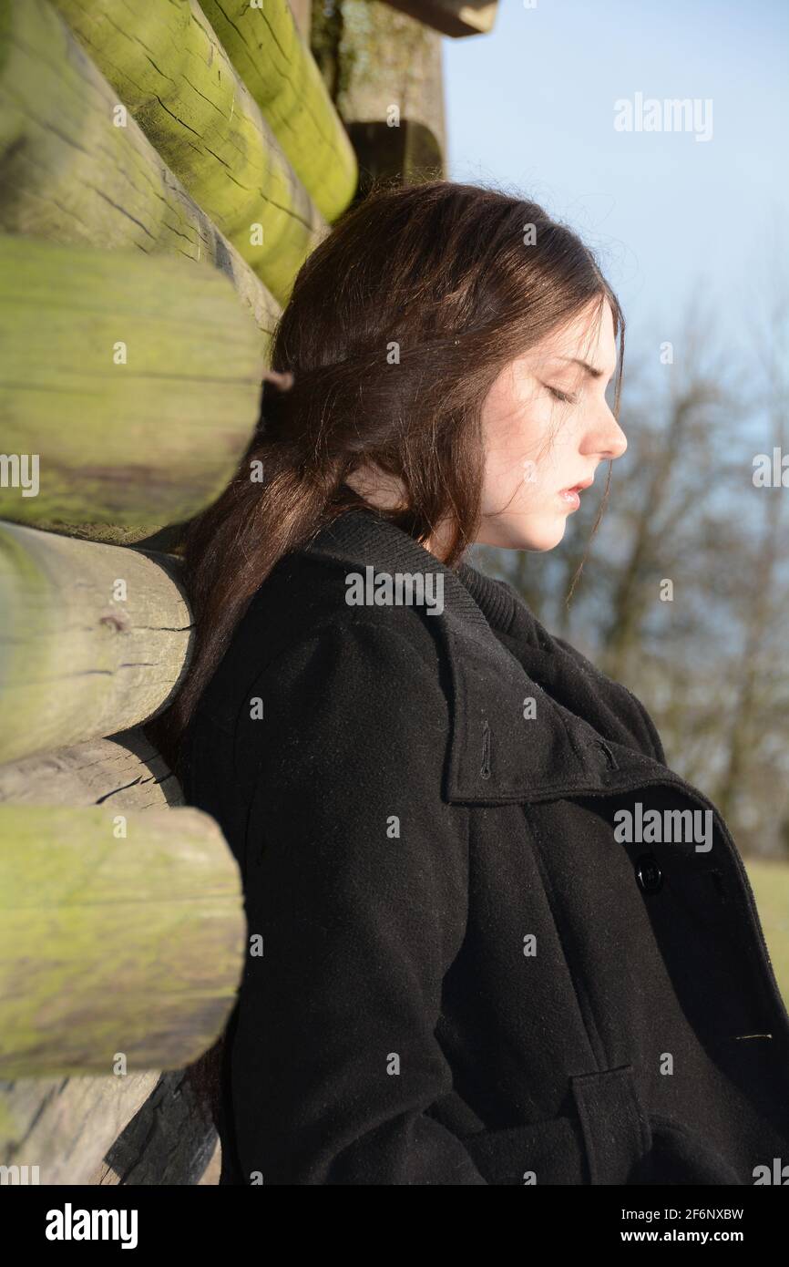 Young woman from the side, with black coat, leaning against a wooden hut, head down, with a serious expression on her face and eyes closed, enjoying t Stock Photo