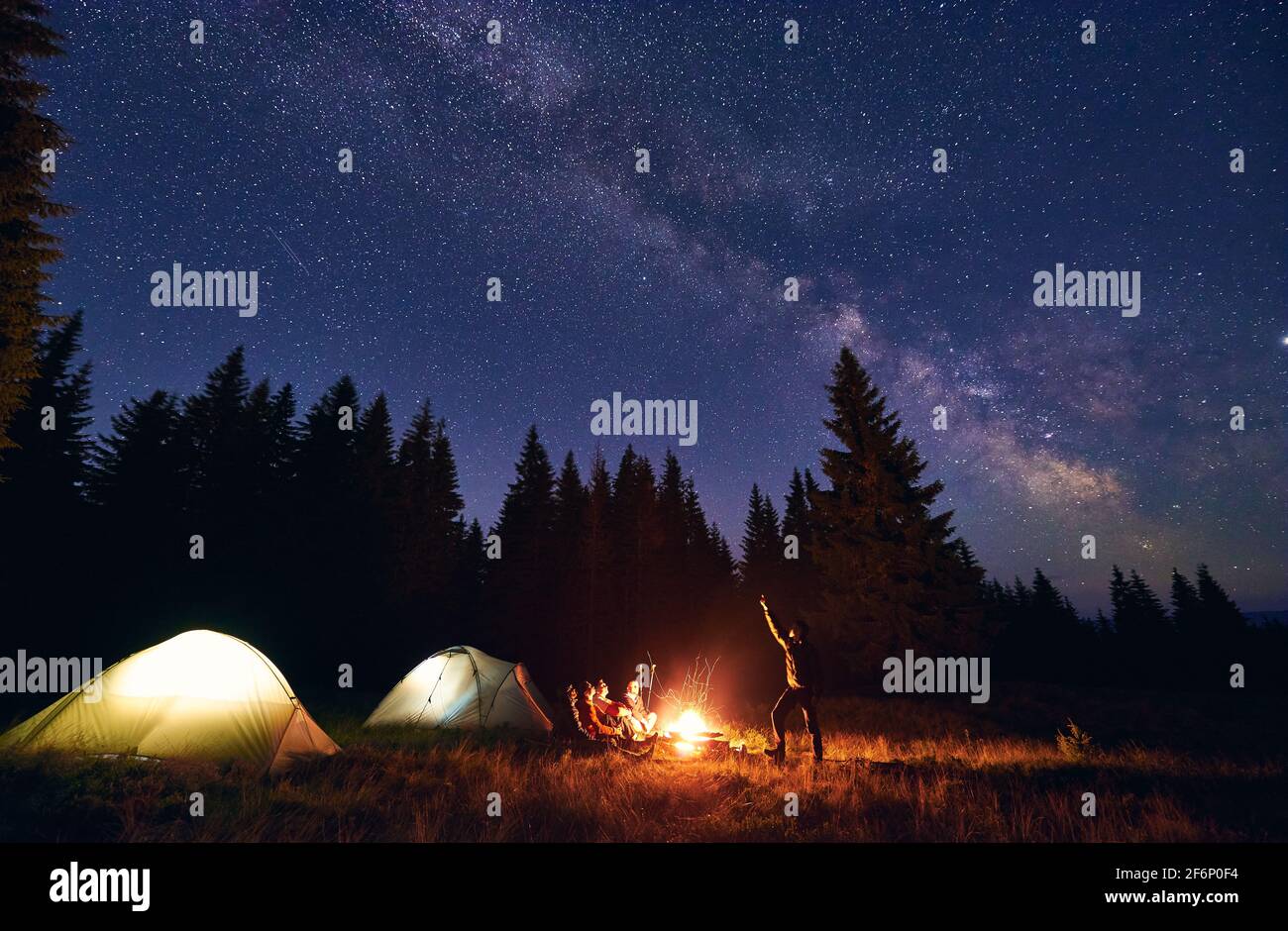 Man is showing his friends Milky Way over tent city. Guys sitting by fire  on the background of spruce forest and enjoying the evening starry sky.  Outdoor recreation. Night camping in the