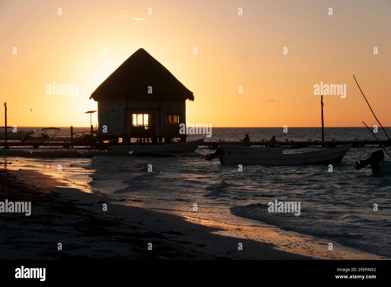 A small fishing shack on a wooden pier over the sea at sunset on Holbox Island in Mexico. In the background are the sky and fishing boats. Travel Tourism Stock Photo