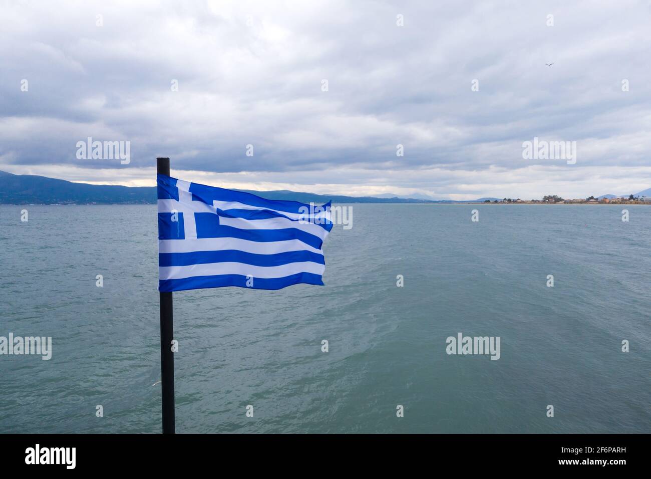Greek flag waving on sea water and cloudy blue sky. Oropos sea Gulf of Evia, Greece. Destination for holiday, travel. Village and mountains for backdr Stock Photo