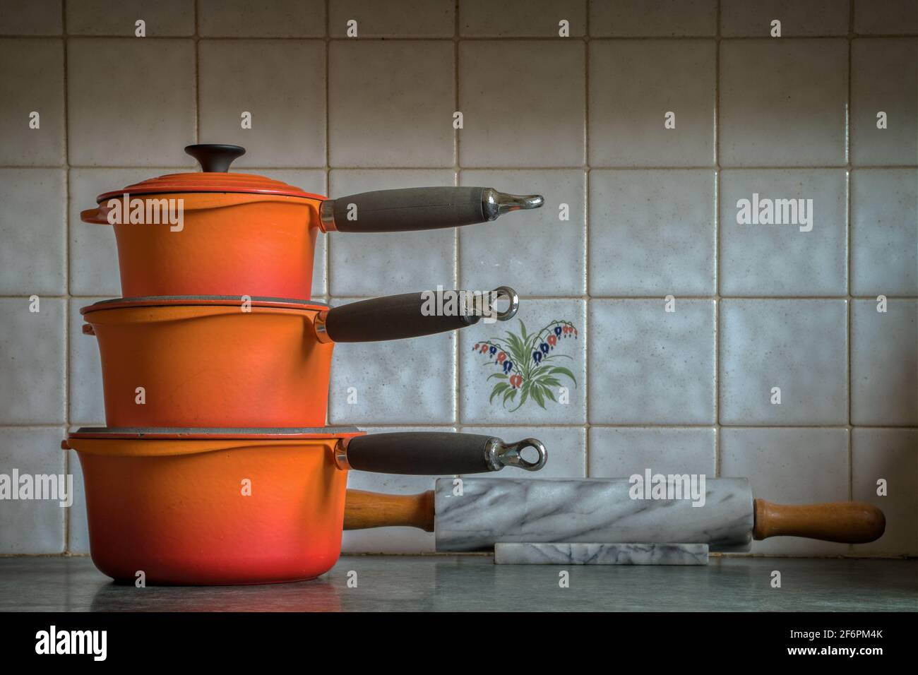 Three orange cast iron saucepans stacked up on a kitchen counter; marble rolling pin; decorated tiles in the background. Stock Photo