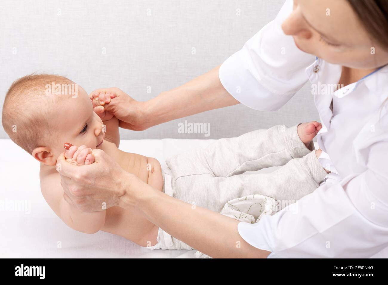 Physiotherapist performing sit-up exercise with six month baby girl in pediatric clinic. Doctor pulling infant up into sitting position to strengthen Stock Photo
