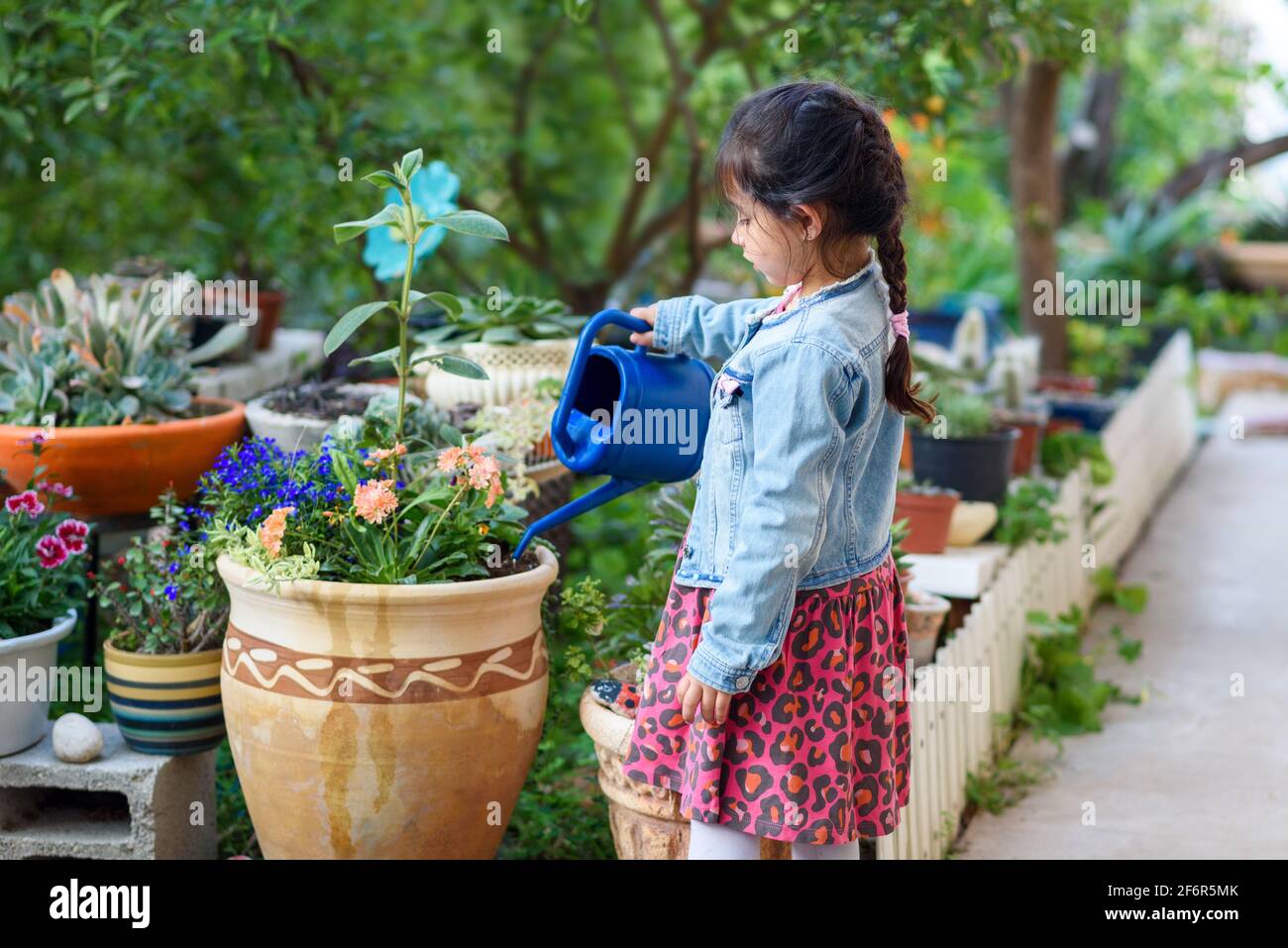 Cute little girl watering flowers in the garden at summer day. Kid helps to care for the plants with a blue watering can in the garden. Stock Photo