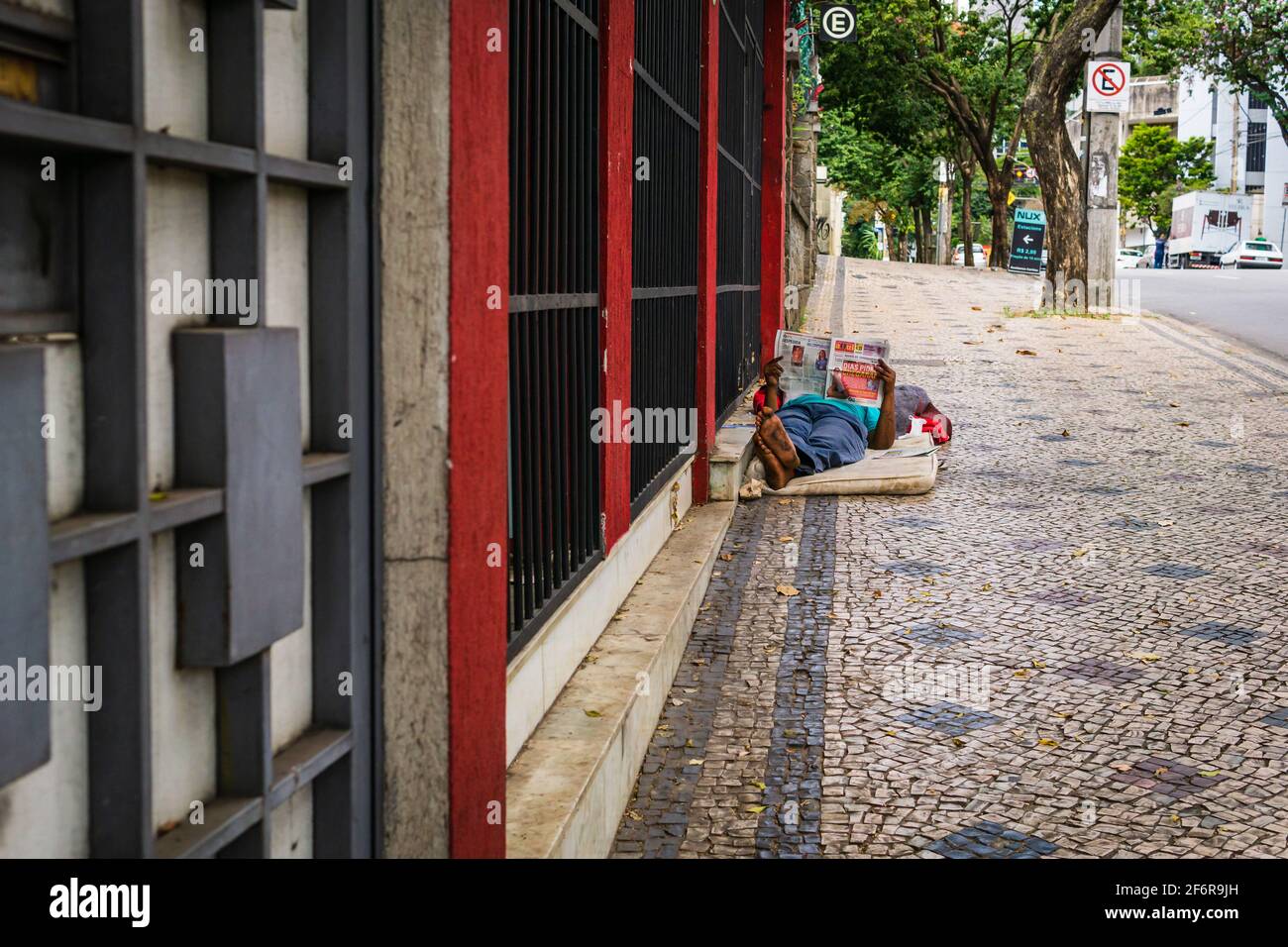 Brazilian homeless man LYING on a mattress on the sidewalk of a street in Belo Horizonte, Brazil. He is reading a newspaper. Stock Photo