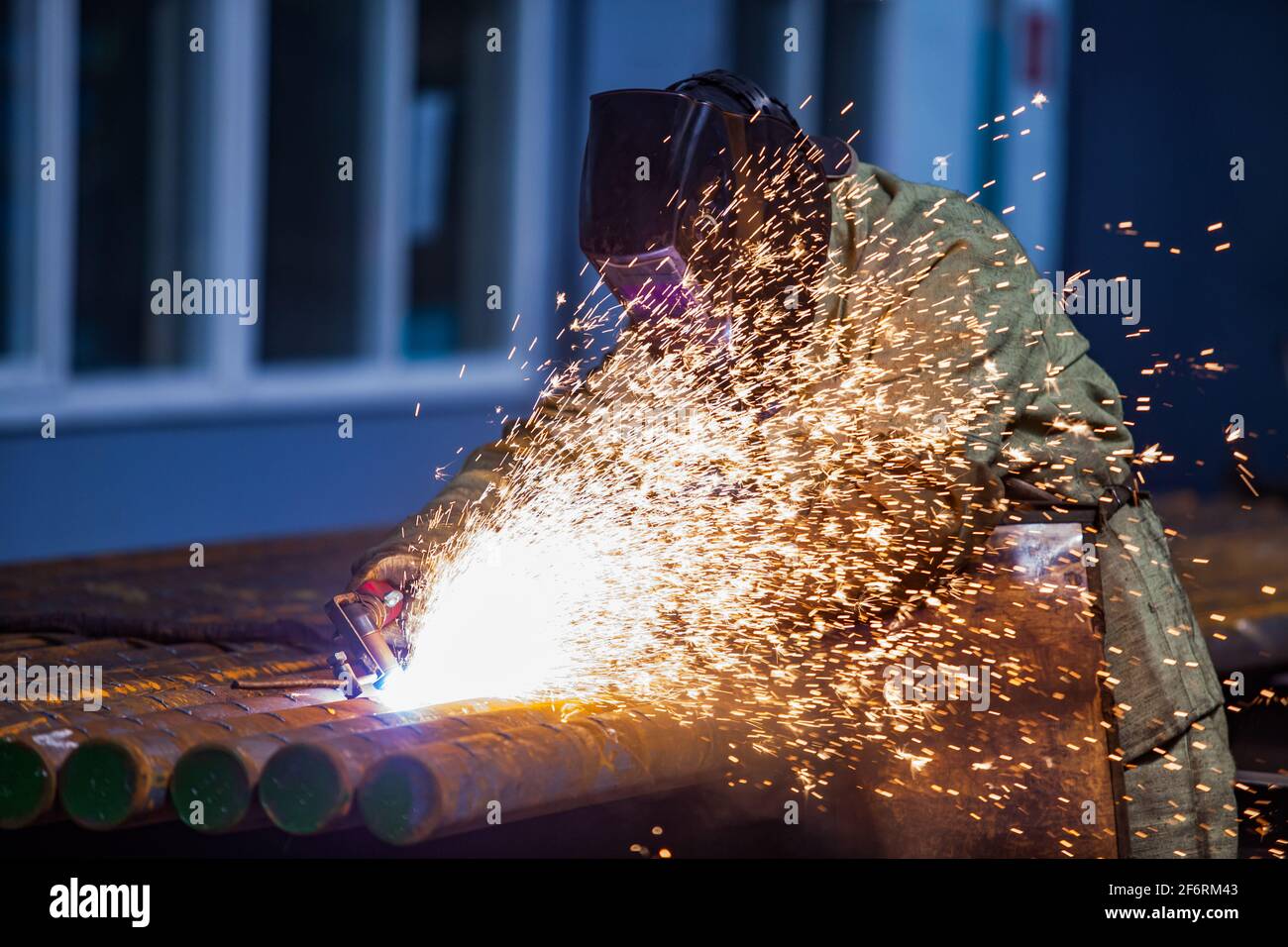 Welder worker in protective mask. Welding steel tubes on manufacturing plant. Sparks and smoke. On blue factory background. Stock Photo