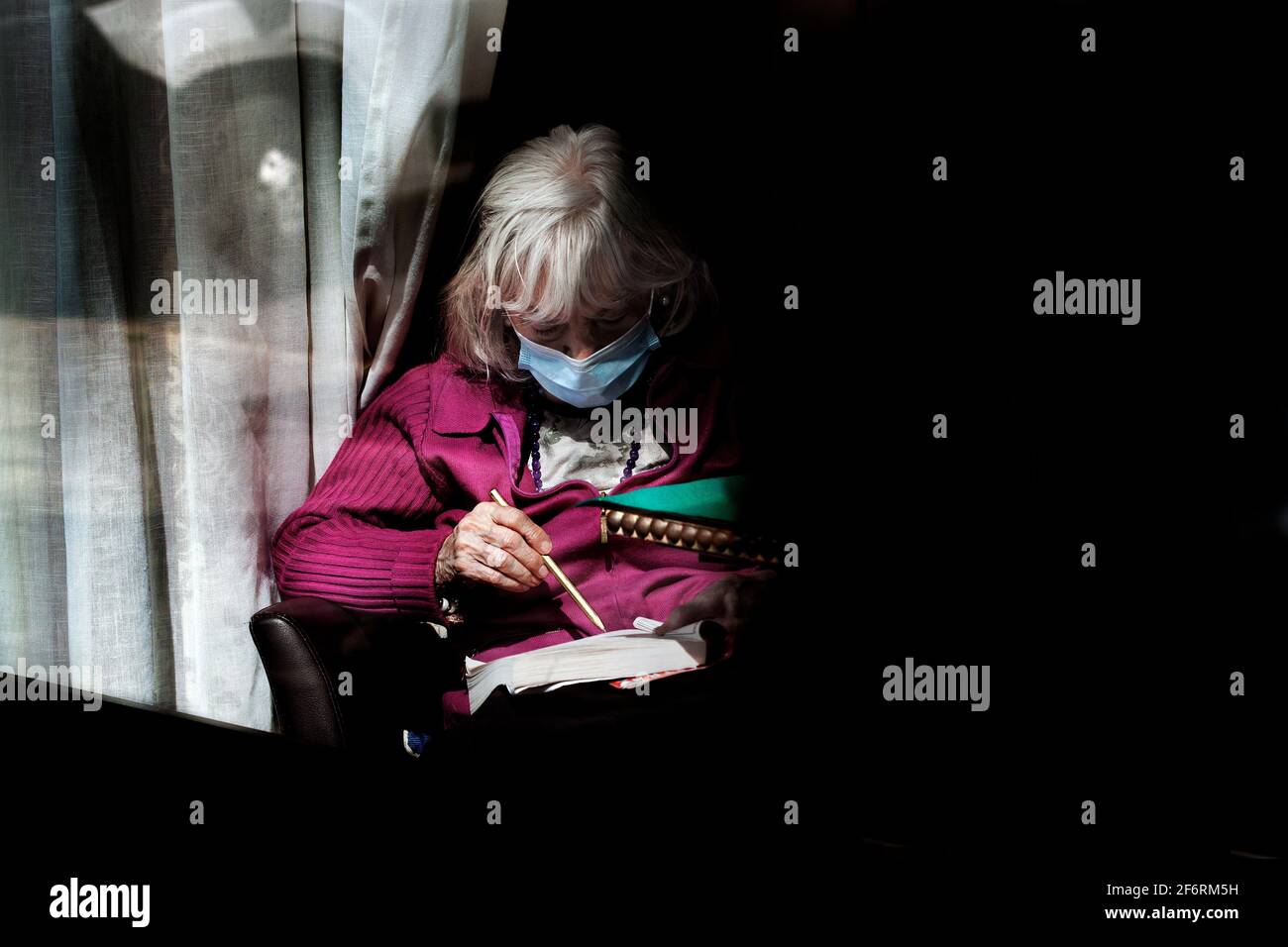 Old woman sat by window of her residence doing a word search puzzle, Barcelona, Spain Stock Photo