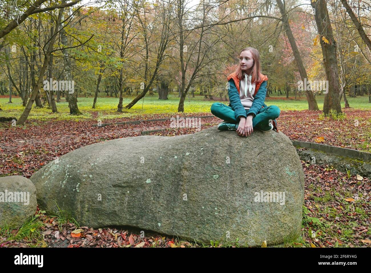 School age girl in turquoise and orange clothes sits on a huge shapeless boulder in an old autumn park, color editing photo Stock Photo