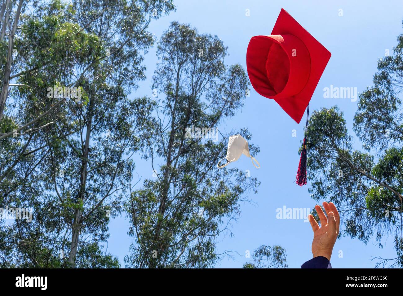 hand of a graduating student throwing his graduation hat and mask into the air over a blue sky on campus. Concept of graduation and social distancing. Stock Photo