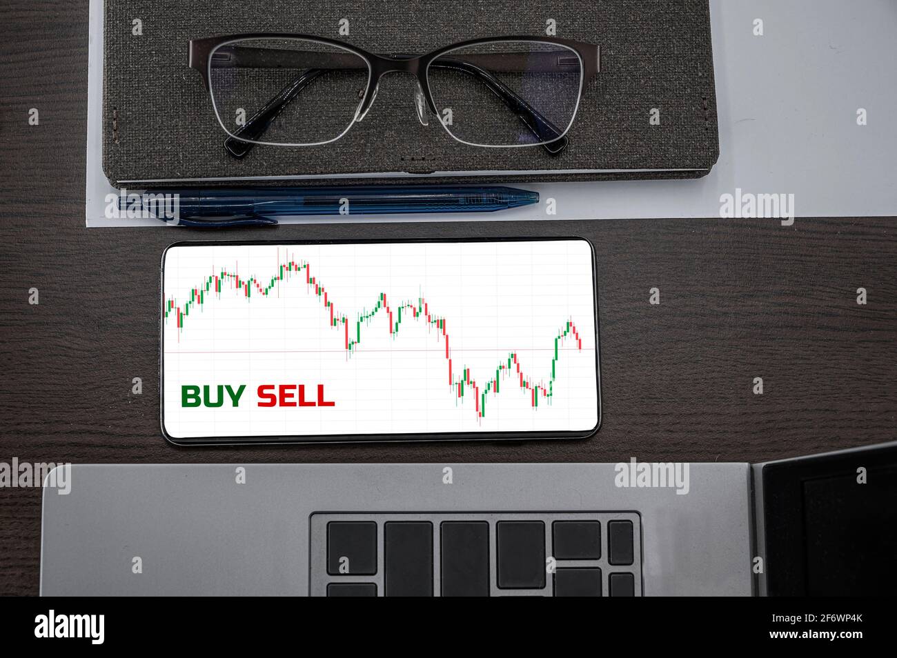 Buy or sell stocks concept. Top view of stocks price candlestick chart in phone on table near laptop, notepad and glasses with inscription buy sell. B Stock Photo