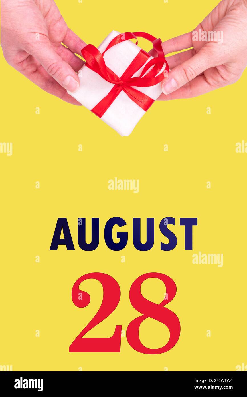 August 28th. Festive Vertical Calendar With Hands Holding White Gift Box With Red Ribbon And Calendar Date 28 August On Illuminating Yellow Background Stock Photo