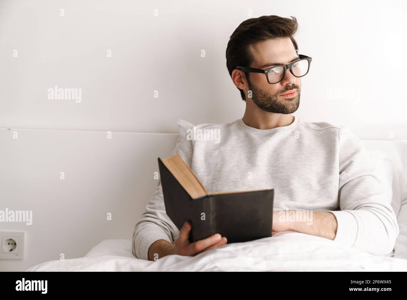Focused young man in eyeglasses reading book while resting in bed at home Stock Photo
