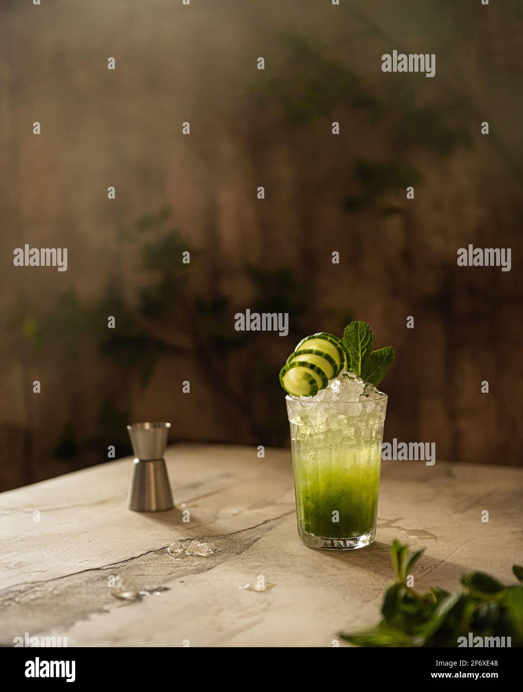 Still life with glass of cold vegetable lemonade with cucumber, mint and crushed ice. Refreshing drink, metal jigger and fresh herbs in natural light. Stock Photo