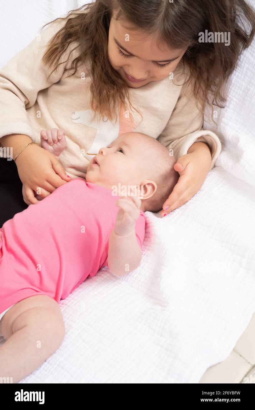 Four year old girl interacting with two month old baby sister, gently touching her head as she looks at her Stock Photo