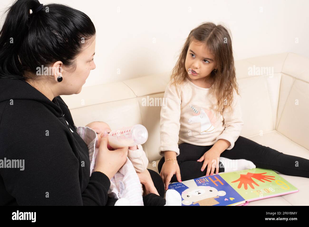 Four year old girl looking at picture book and talking with mother, who is feeding 2 month old baby sister Stock Photo