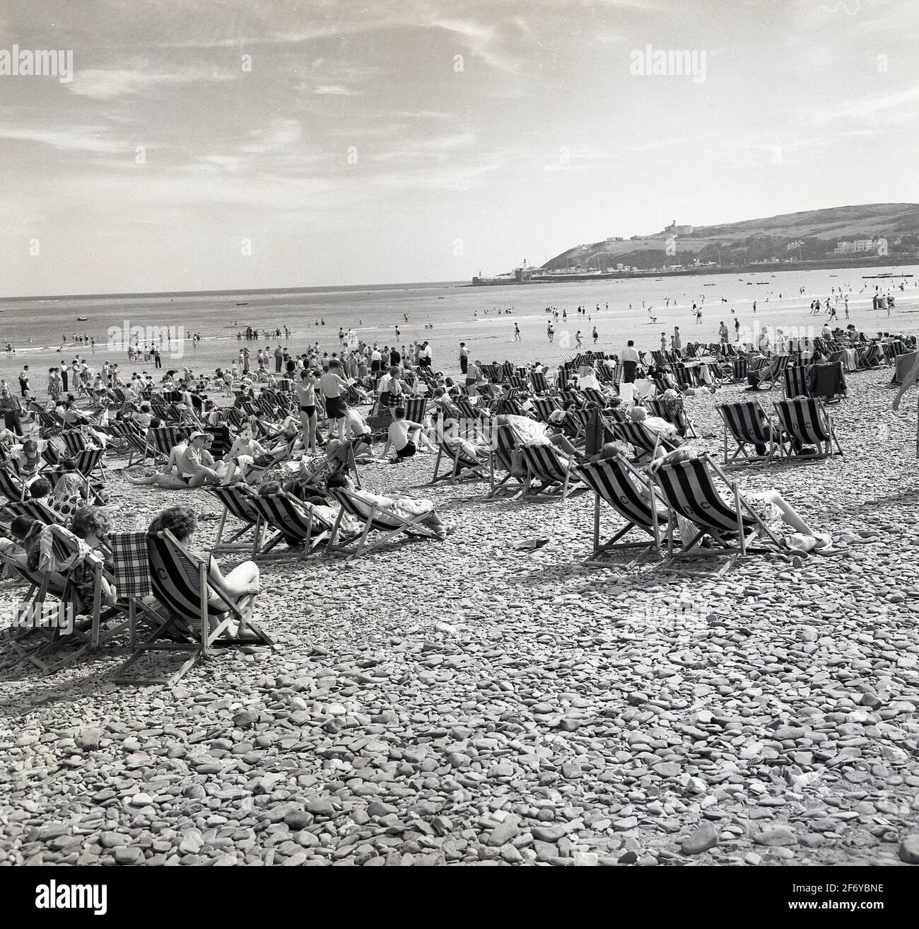 1950s, historical, at the coast, holidaymakers sitting in deckchairs on a stoney beach, England, UK. The simple deckchair is an icon of the British seaside and and relaxing in one on a summer's day is a tradition dating back to Victorian times. In post-war Britain, before the advent of overseas holidays, it remained as popular as ever, with the press reporting that on one day in August 1957 over 66,000 deckchairs were hired out on Blackpool beach. While those numbers may not be seen again, the deckchair has an evergreen appeal, based on nostalgia and its simple, effective design. Stock Photo