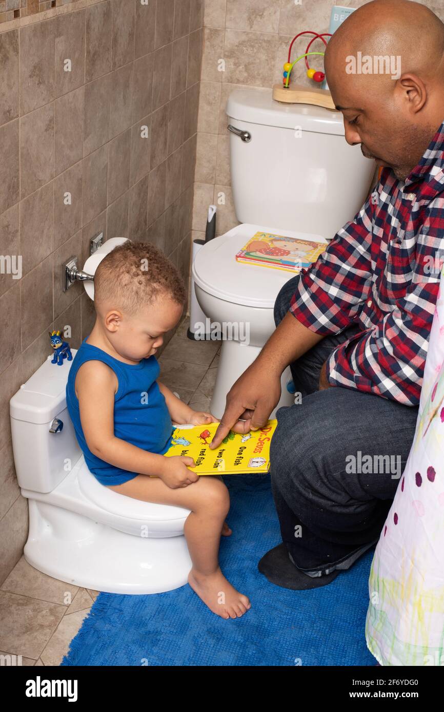 Toilet training: 2 year old boy sitting on potty in bathroom, looking at books with father Stock Photo