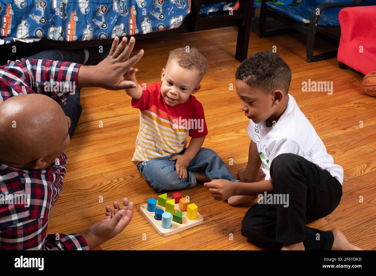 Brothers, ages 2 and 6 playing with geometric shape sorter, father giving 2 year old a “high 5” for success Stock Photo