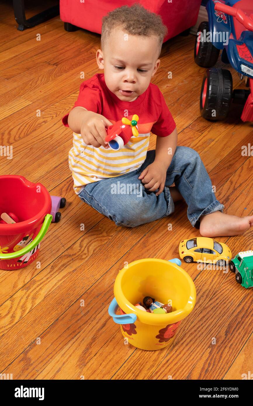 2 year old boy sitting on floor playing with small people and vehicles in two color full plastic buckets, making sounds as he makes airplane fly Stock Photo