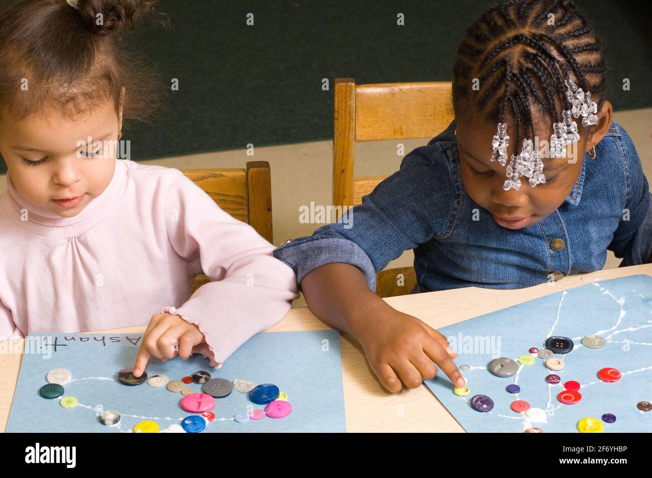 Education preschool 3-4 year olds two girls sitting side by side doing art activity gluing buttons onto construction paper, one using right hand and t Stock Photo