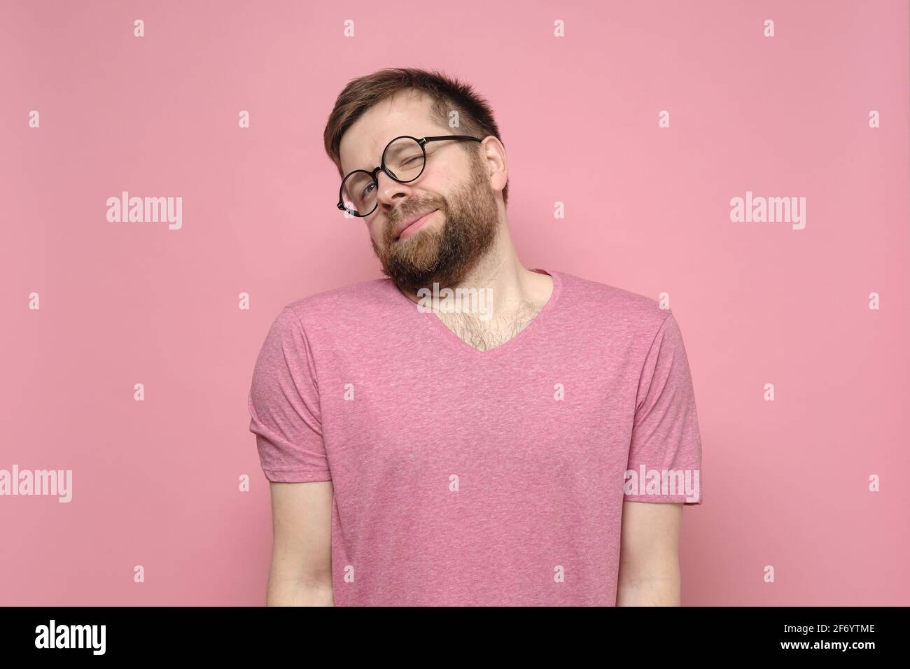 Cute, shy man with glasses hunched shoulders and looked with narrowed eyes at the camera. Pink background. Stock Photo