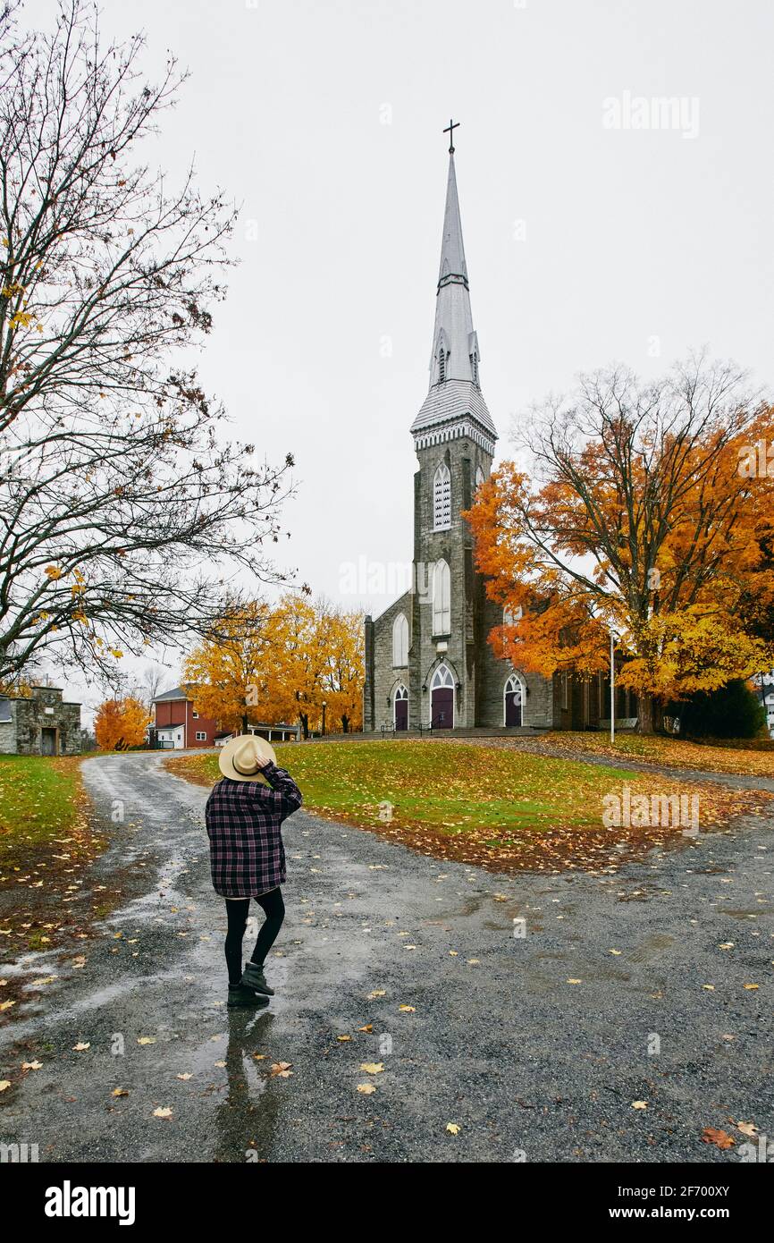 Back view of anonymous female in checkered shirt touching hat and admiring old church on gray autumn day in town Stock Photo