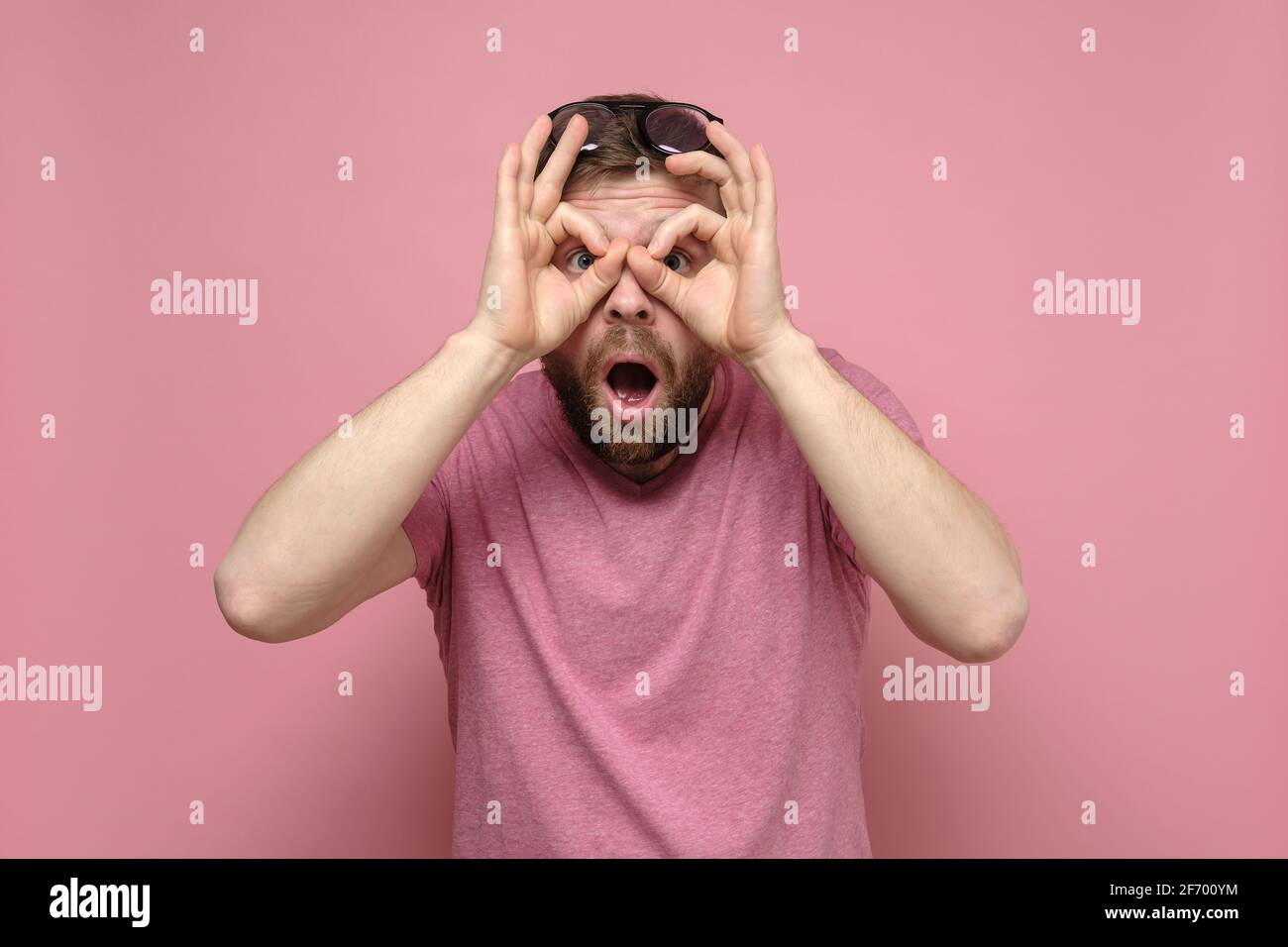 Bizarre, shocked man put fingers to eyes like binoculars and looks through them with mouth open in surprise. Pink background. Stock Photo