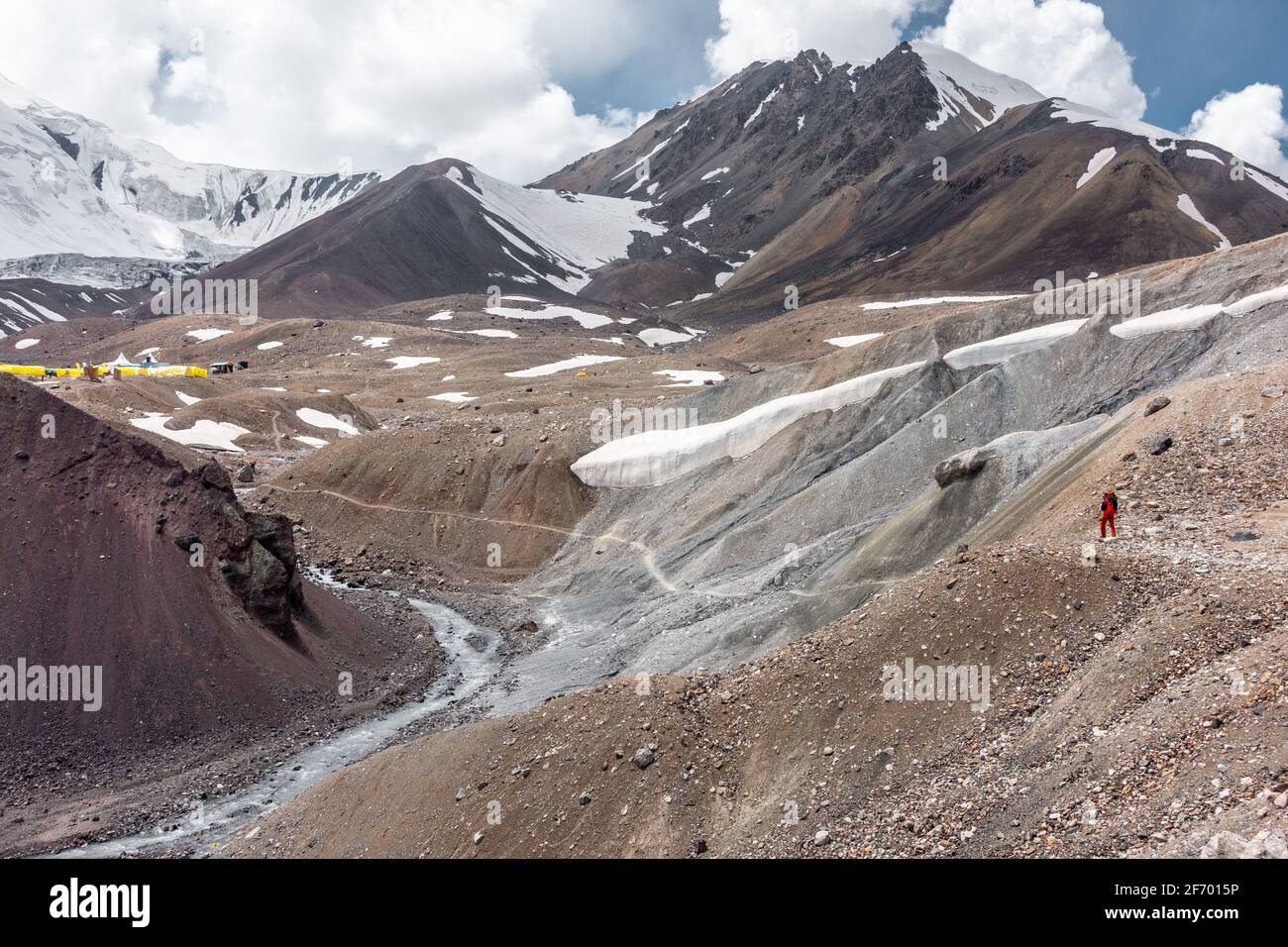 Adult man with a large backpack approaching the base camp in Pamir mountain range with enormous snowcapped Lenin Peak in distance, Kyrgyzstan Stock Photo