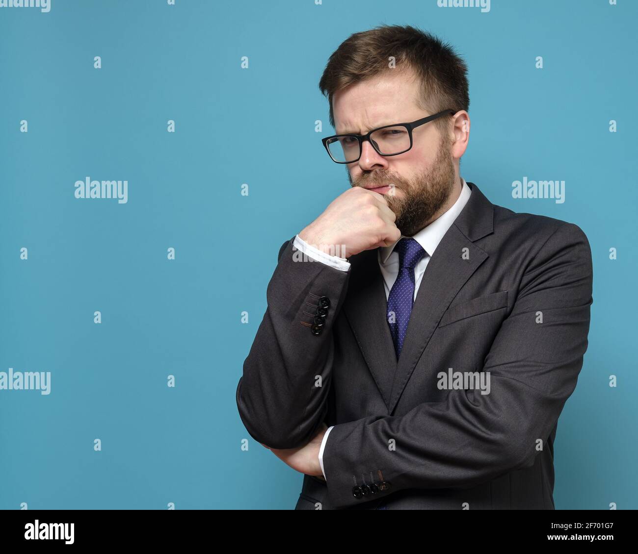 Suspicious, pensive business man in a suit and glasses looks sternly, holding hand to his face. Copy space. Blue background. Stock Photo